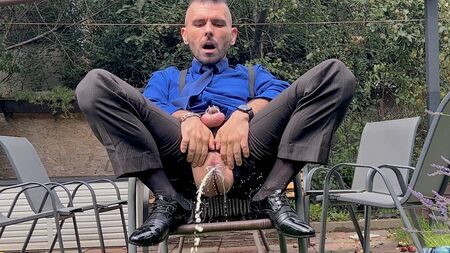 スーツ姿の男が、ペニスを拘束された状態で、男のアナルをいじりながら、透けるソックスを履いたまま人前で放尿している